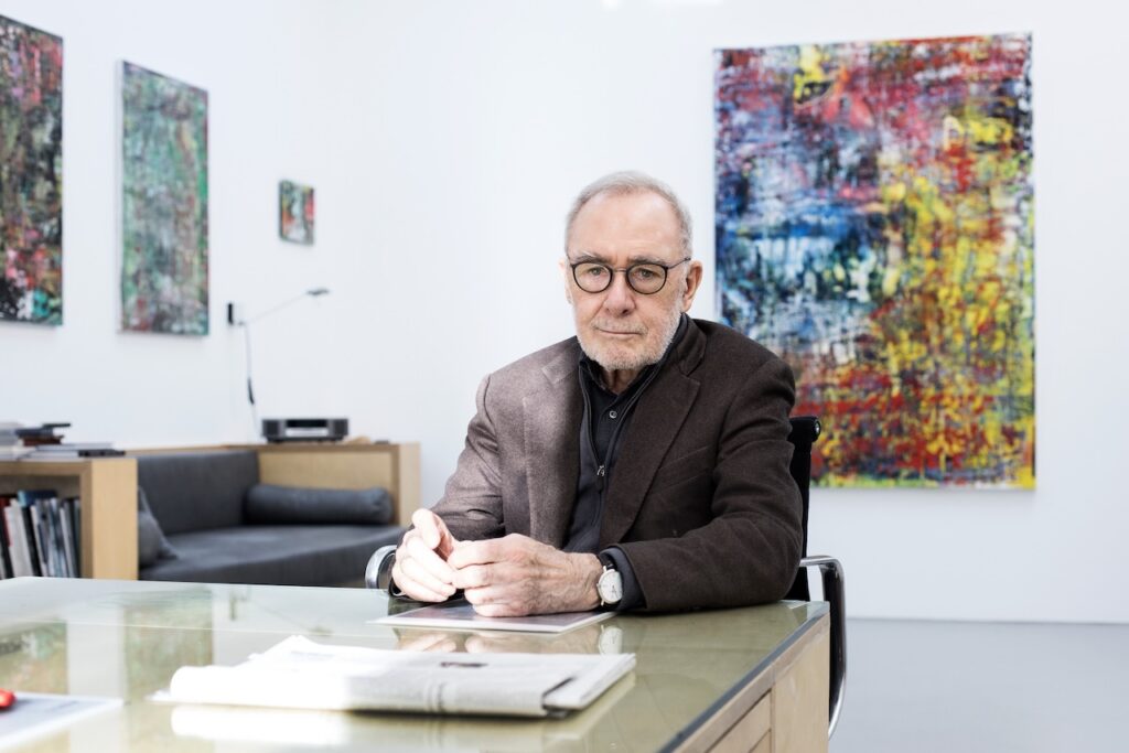 Gerhard Richter sits at a desk in a gallery with colorful abstract paintings on the walls, wearing glasses and a blazer.
