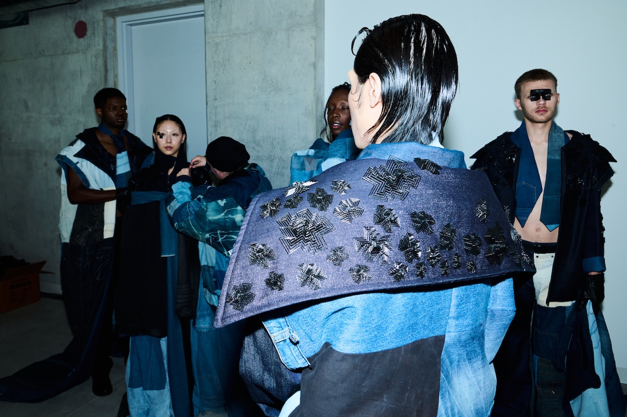 Models wear layered denim outfits with dark embellishments backstage as a stylist adjusts a model’s clothing.