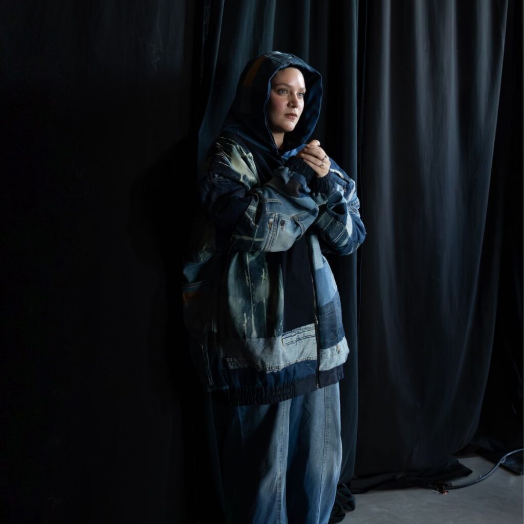 A person wears a Roni Helou patchwork denim hooded jacket and matching denim jeans while standing against a black curtain.