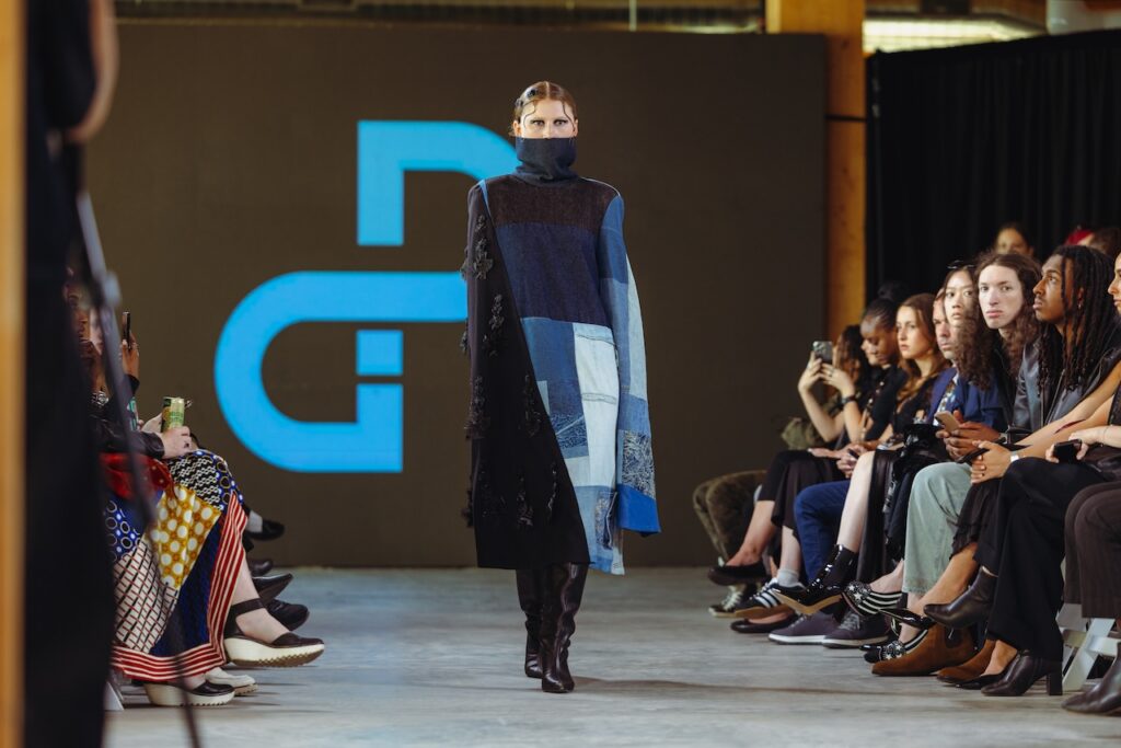 A model wears a black and blue patchwork coat with a high neck while walking down a runway in front of a seated audience.